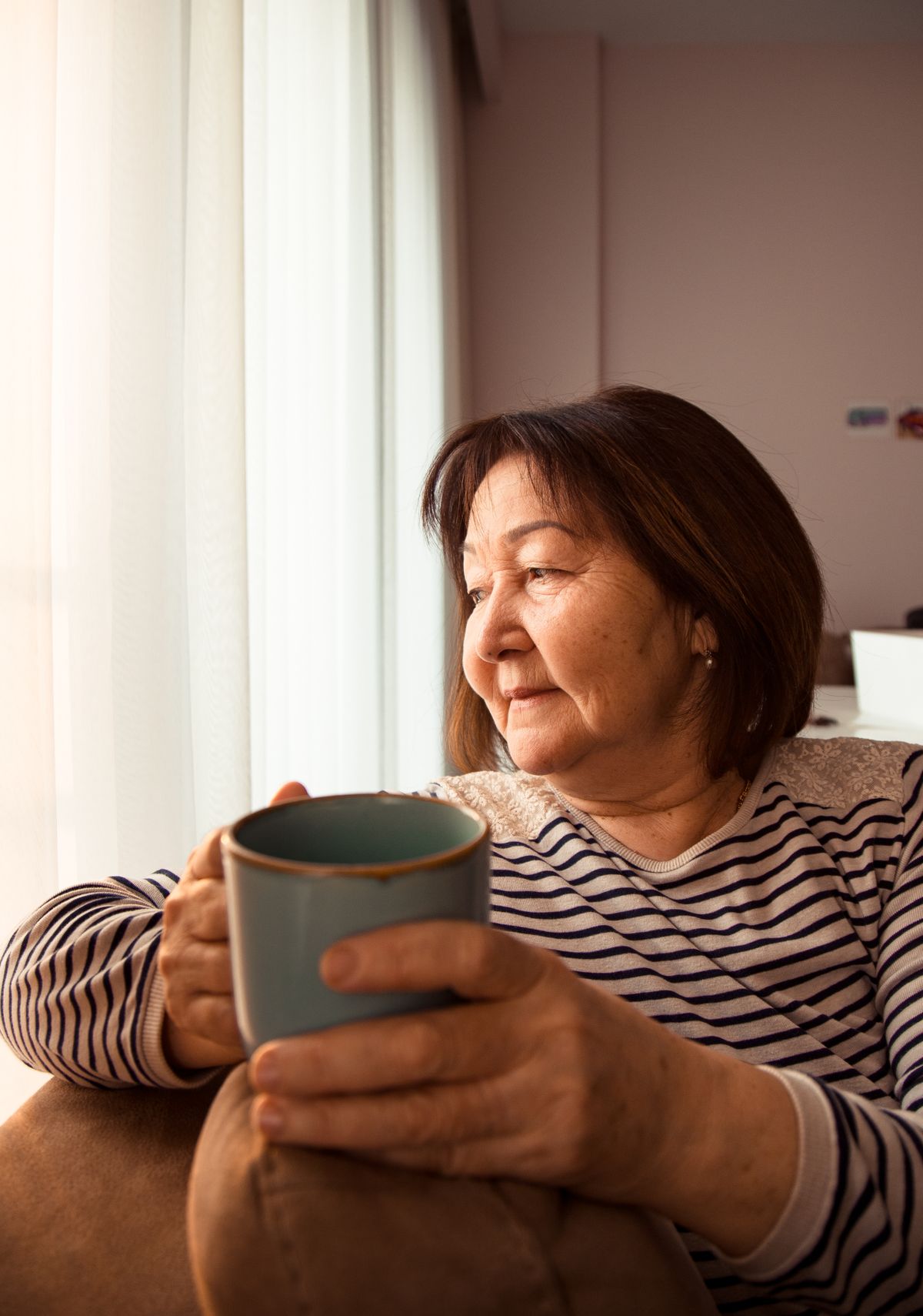 An older woman smiling with a hot drink while relaxing by a window at home.